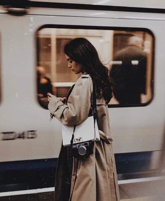Woman in a trench coat on a train platform, holding a phone and camera, with a train moving in the background.