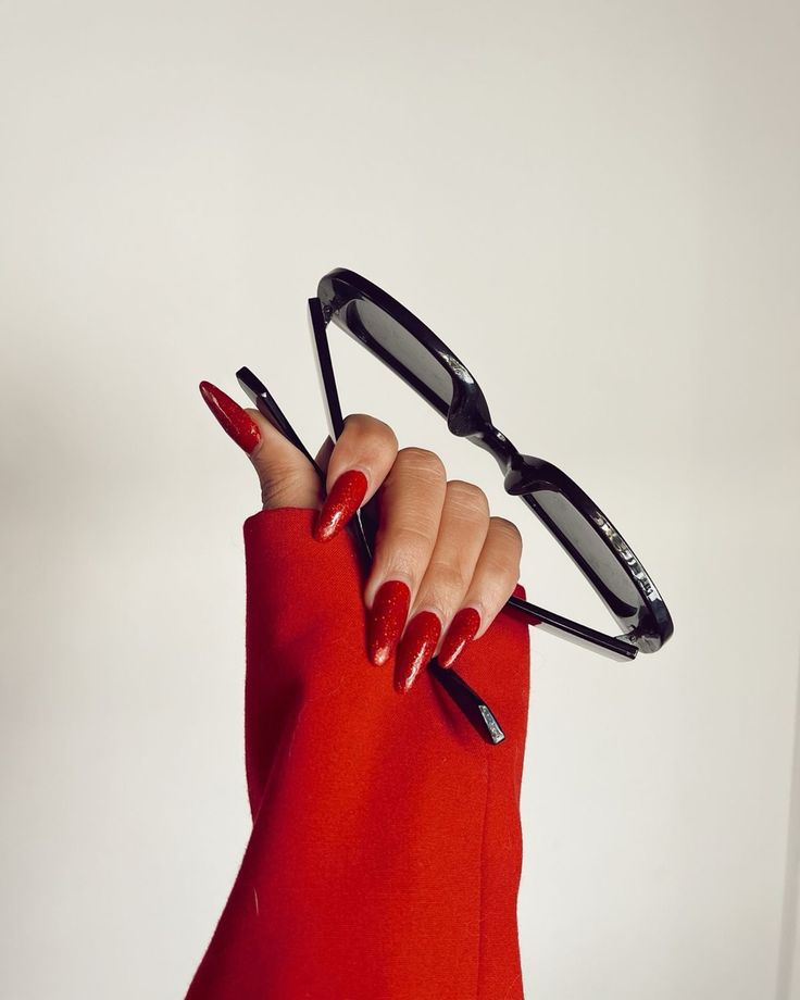 Hand with red nails holding sunglasses, dressed in red sleeve against white background. Fashion and style concept.