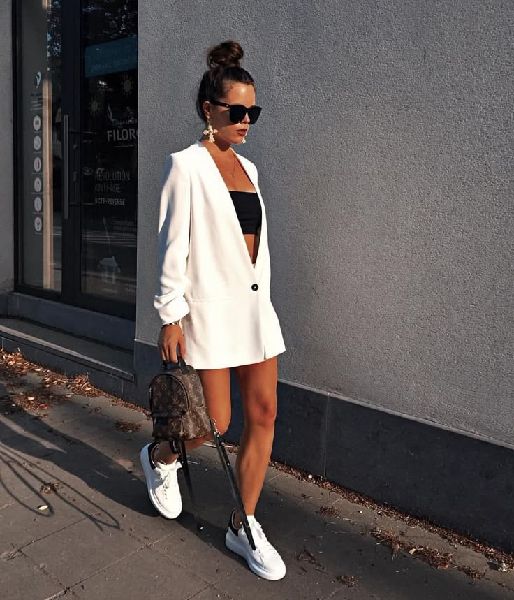 Fashionable woman in white blazer dress, sunglasses, and sneakers, carrying a small backpack on city street.