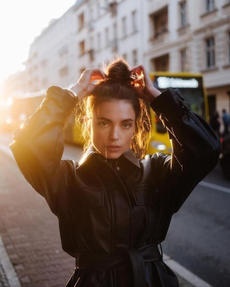 Woman in black jacket adjusts hair in sunlit street with yellow buses in the background.