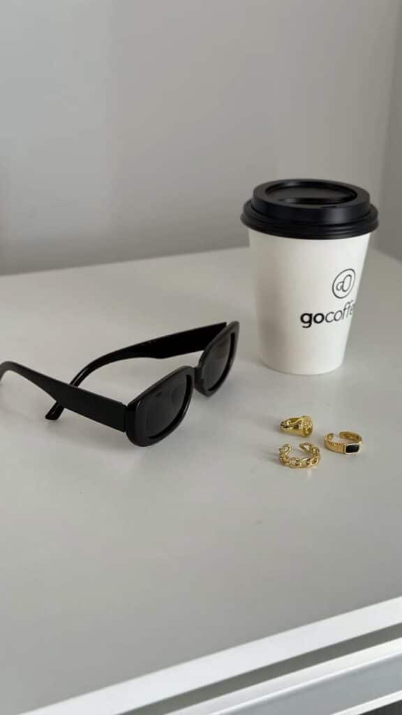 Sunglasses and gold rings beside a coffee cup on a white surface, minimalist aesthetic.