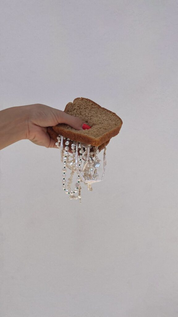 Hand holding a slice of bread with hanging pearl jewelry against a plain background.