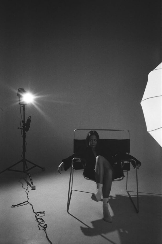 Black and white photo of a woman sitting on a chair in a studio, with bright lighting and umbrella reflector.