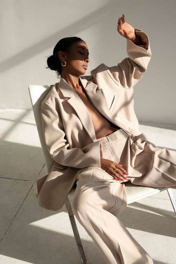 Person lounging in beige suit, hand shielding face from sunlight, casting dramatic shadows in minimalistic setting.