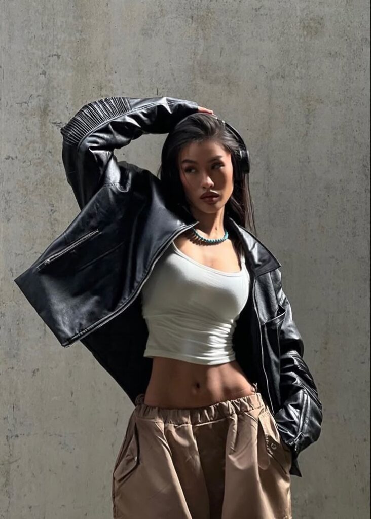 Woman posing in leather jacket and beige pants against a textured wall, showcasing trendy fashion style.