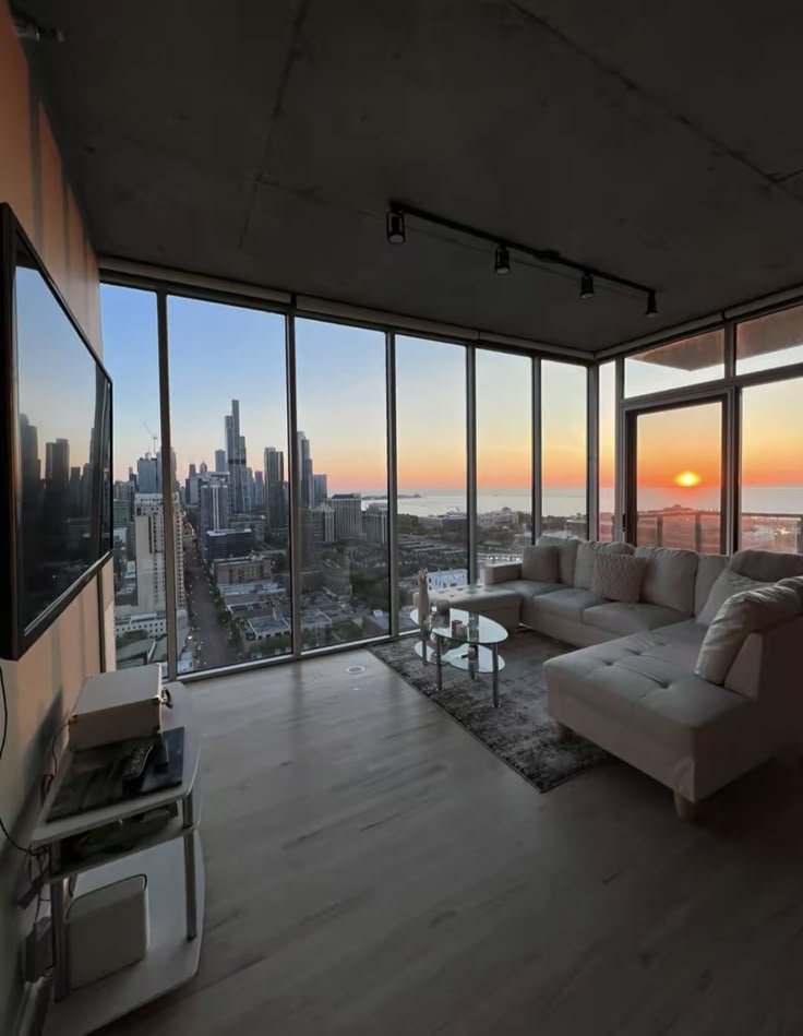 Modern living room with panoramic city skyline view at sunset through large windows.