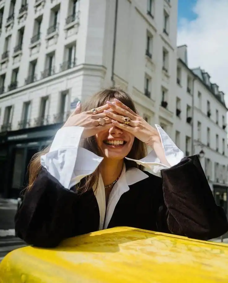 Woman playfully covering face with hands, wearing rings, smiling on city street. Bright sunny day, urban background.
