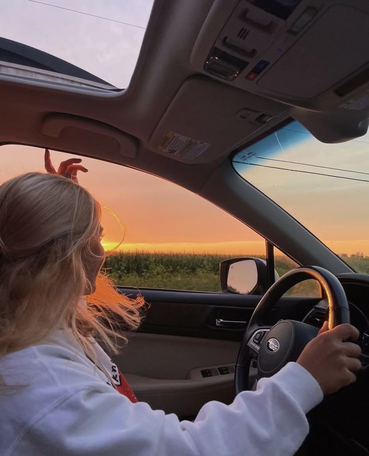 Woman driving car at sunset with open sunroof, enjoying scenic view of fields and colorful sky.
