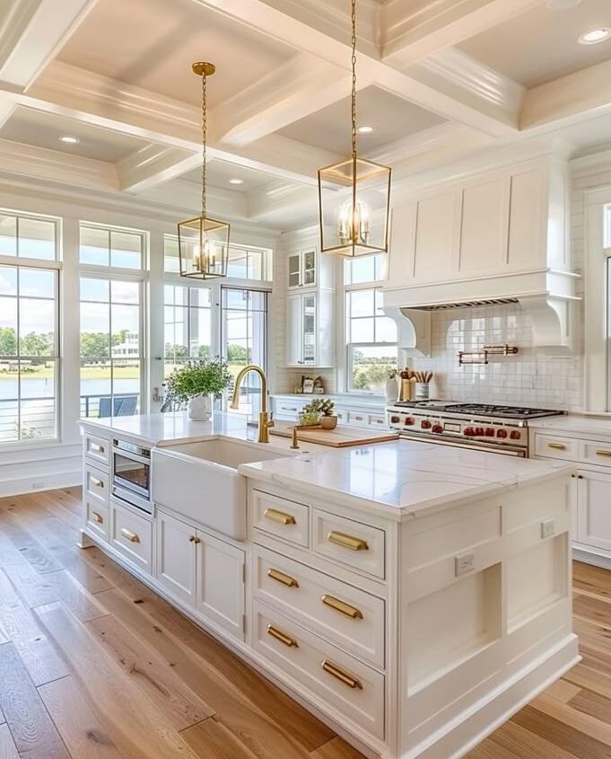 Spacious modern kitchen with white cabinets, gold fixtures, large island, and outdoor views through large windows.