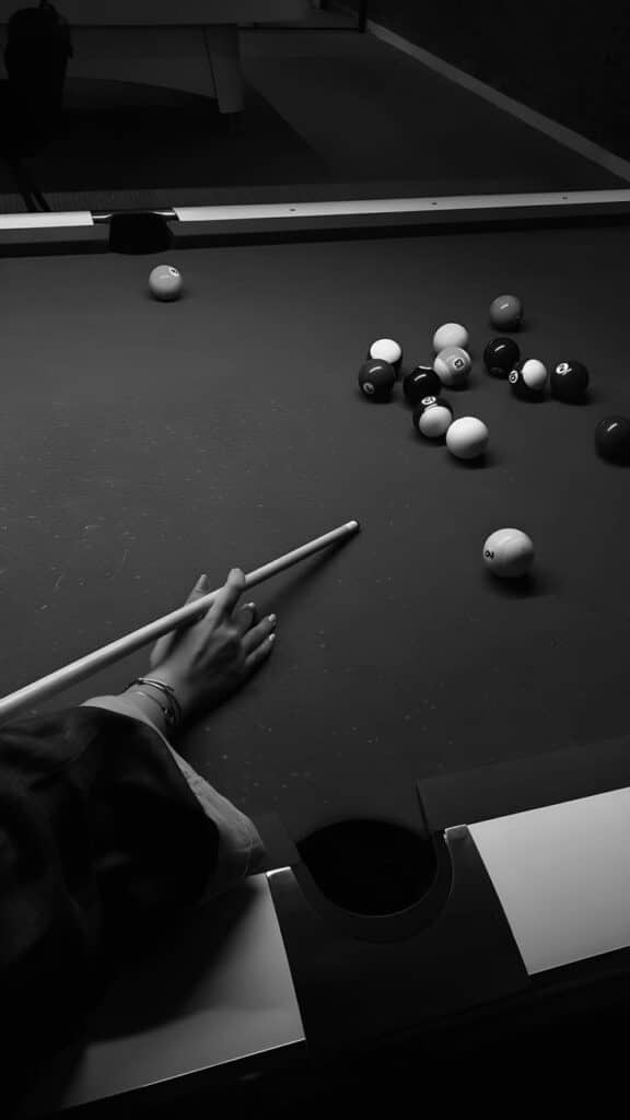 Hand aiming with cue stick on pool table for a shot, surrounded by pool balls in monochrome.