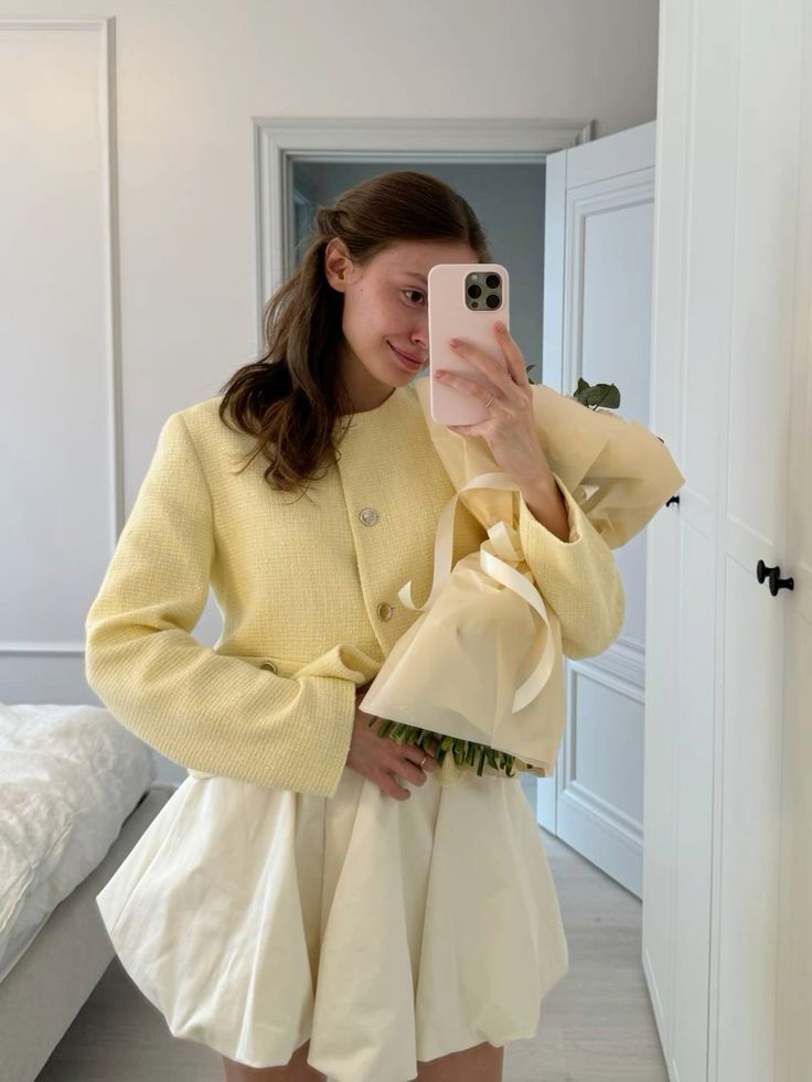 Person taking a mirror selfie holding flowers, wearing a yellow jacket and white skirt in a bright room.