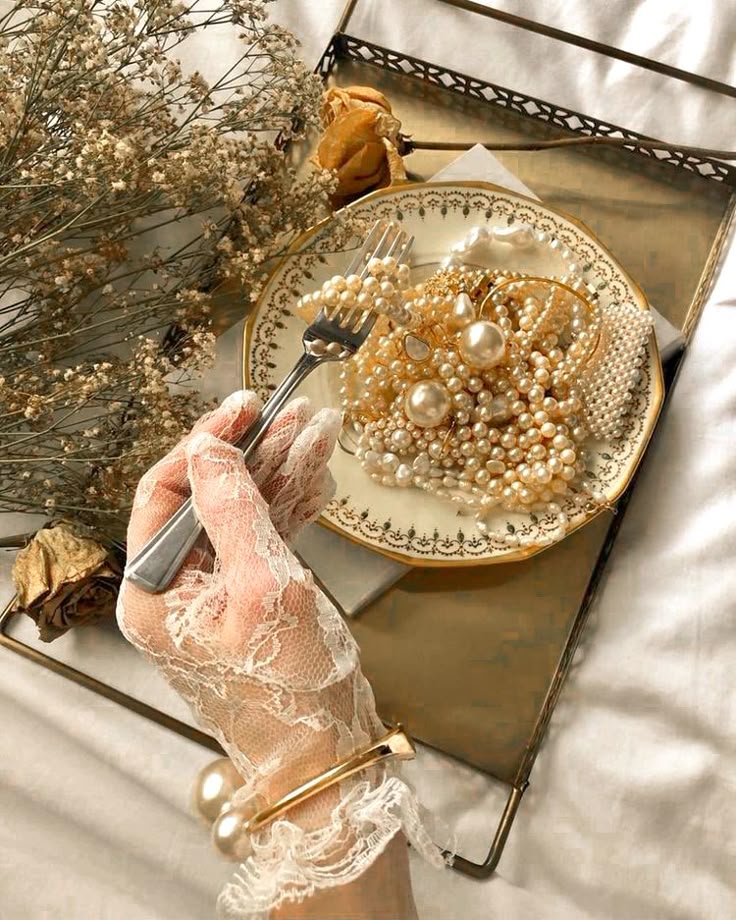 Lace-gloved hand holds fork with pearl jewelry on ornate plate, elegant vintage aesthetic on tray with flowers.