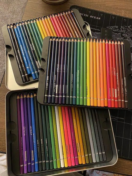 Colorful pencil sets in open metal tins on a table, arranged by hues from reds to blues and greens to purples.