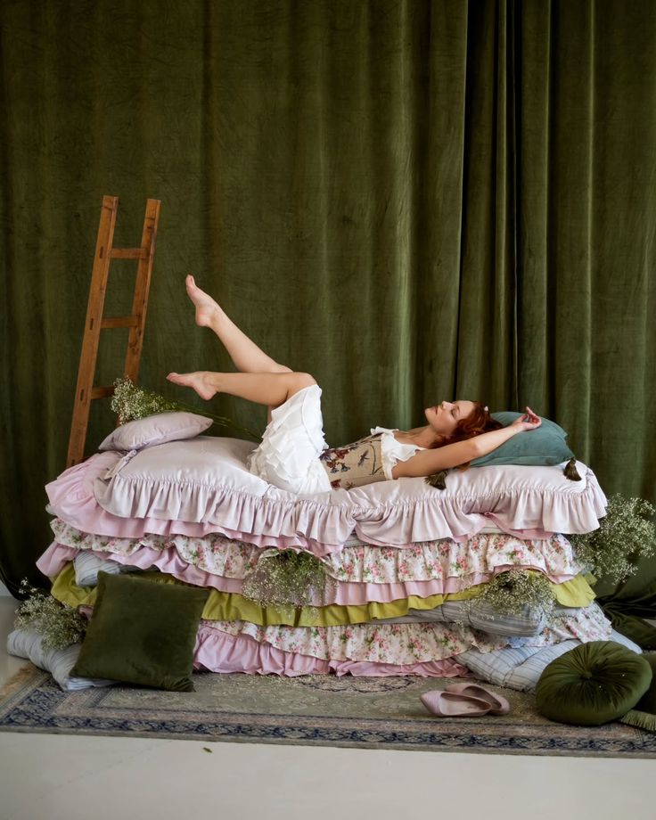 Woman relaxing on layered pillows with vintage decor and green curtain backdrop.