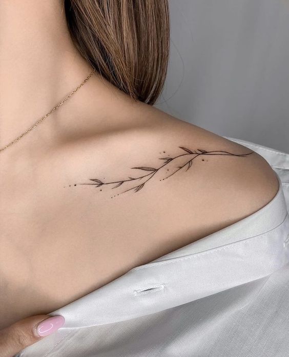 Minimalistic floral tattoo on a woman's shoulder with simple leaves and dots, wearing a white shirt.