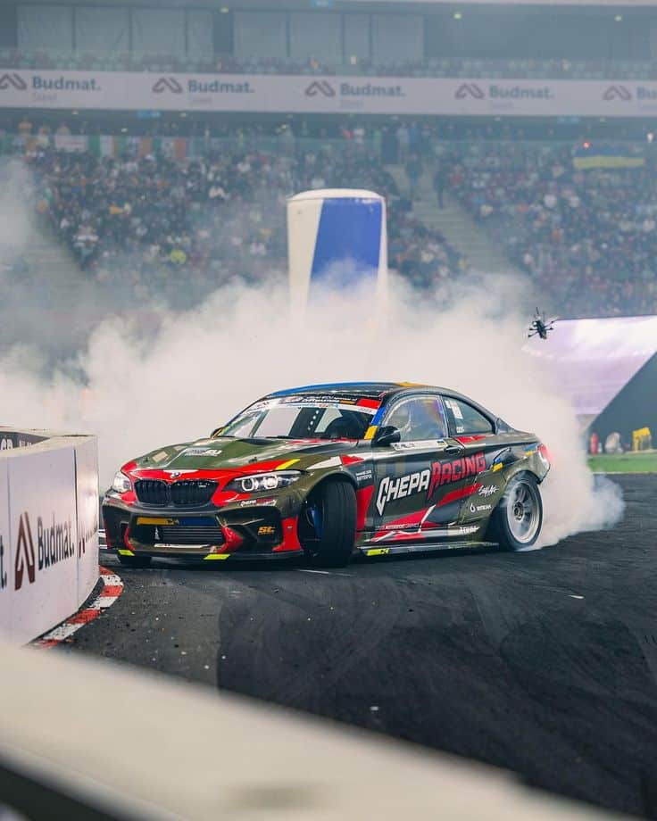 Racing car drifting on a track with smoke during a lively motorsport event, surrounded by a cheering crowd.