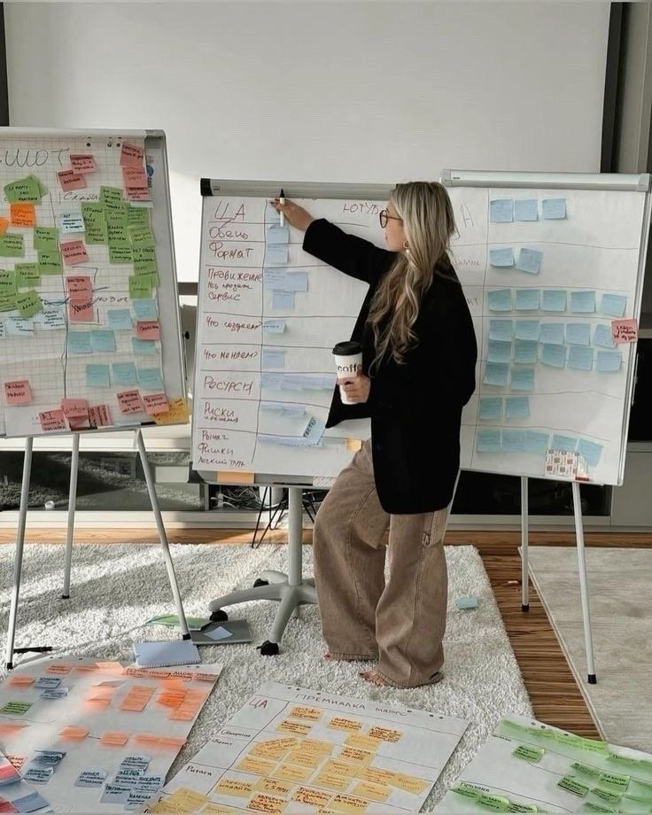 Woman presenting project plans on flip charts, holding coffee. Colorful sticky notes and charts on the floor and boards.