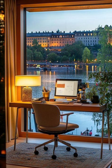 Cozy home office with lake view, featuring a desk, lamp, chair, computer, and scenic window overlooking buildings and water.