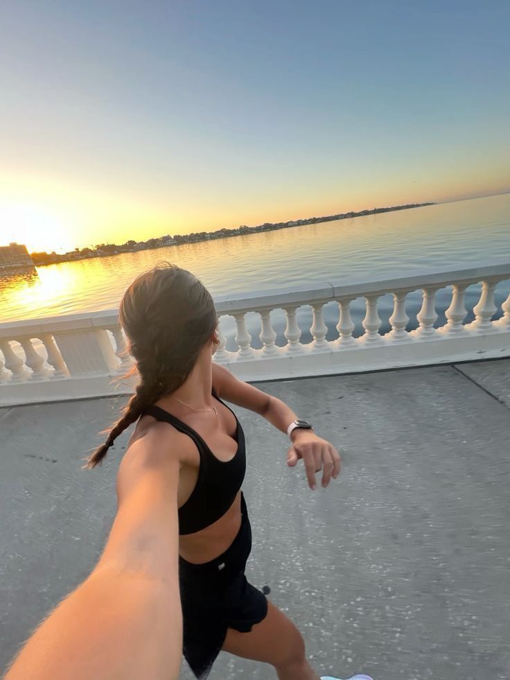 Woman jogging by the waterfront at sunrise, wearing sportswear and tracking fitness on a smart device.