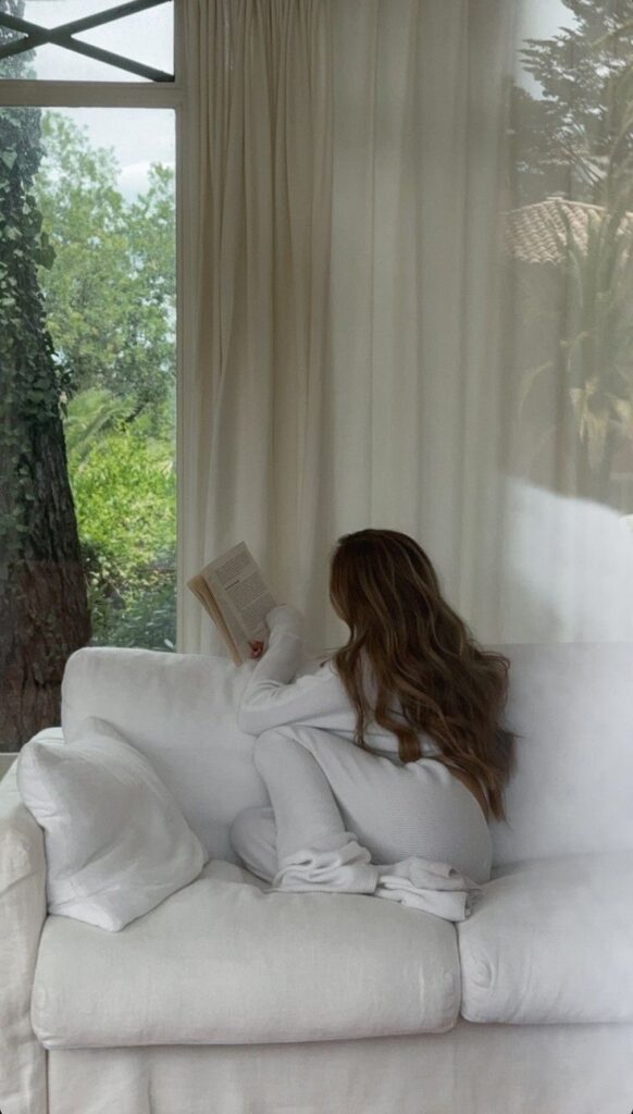 Woman reading on a cozy white couch by a window, surrounded by trees.