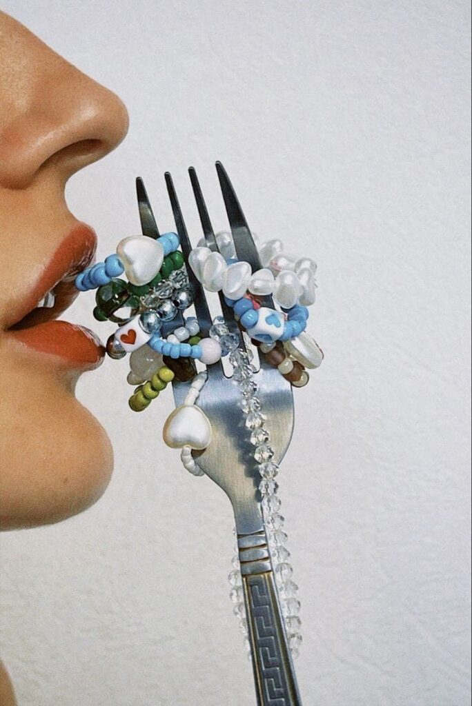 Close-up of a person holding a fork with colorful beaded jewelry, near their mouth, against a white background.