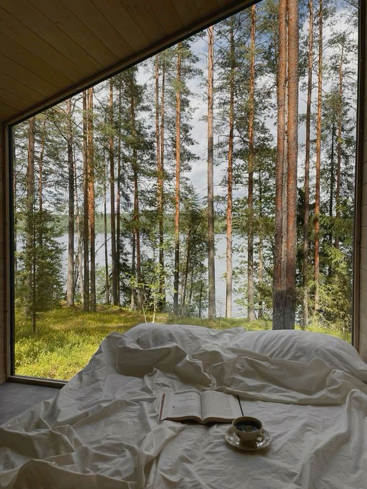 Cozy bedroom with forest view through large window, showcasing bed with book and coffee, creating a serene retreat.