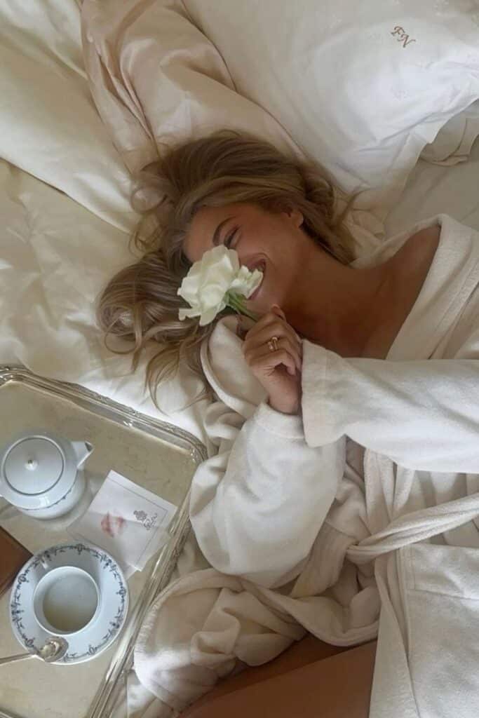 Woman in a plush robe smiling with a flower, beside a breakfast tray with tea set on a cozy bed.
