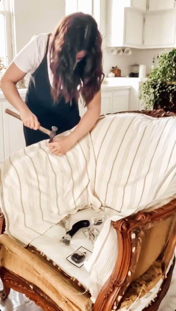 Woman reupholstering vintage chair in bright kitchen with tools like hammer and tape measure.
