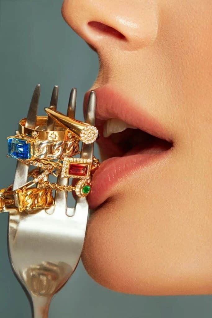 Close-up of lips touching a fork adorned with colorful gemstone rings and gold jewelry.