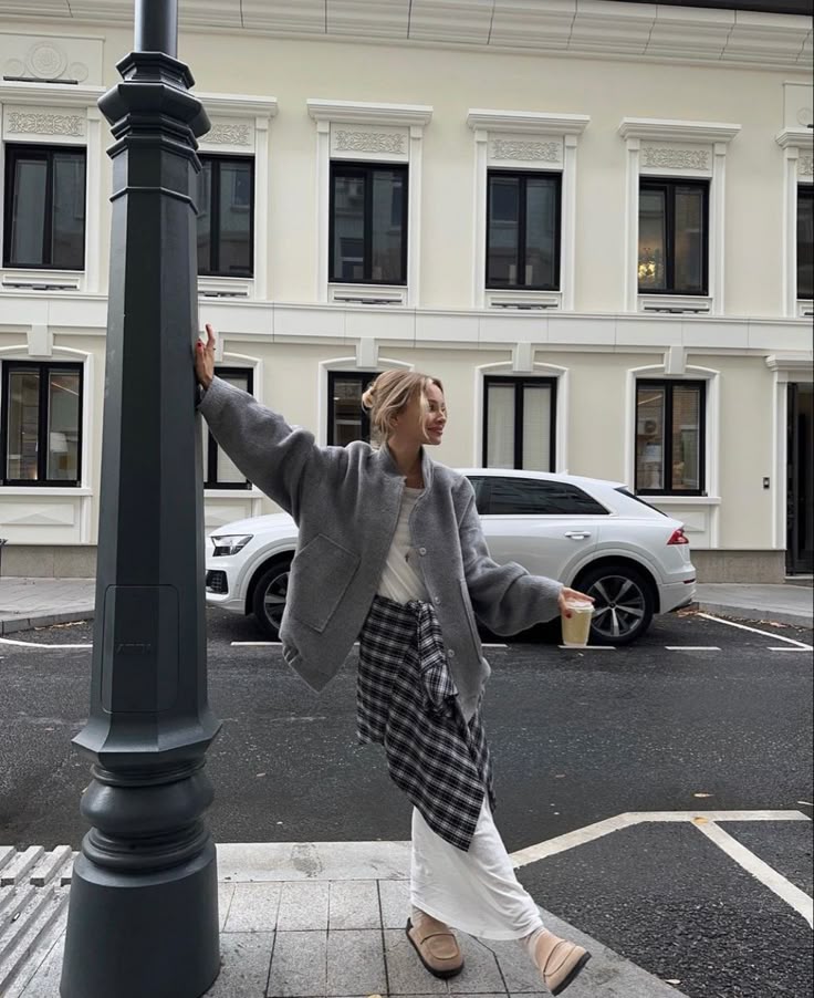 Stylish woman in gray coat holding drink, leaning on lamppost, urban background with white building and parked car.