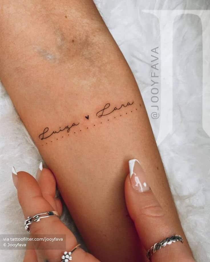 Minimalist tattoo with names and heart on forearm, adorned by manicured nails.