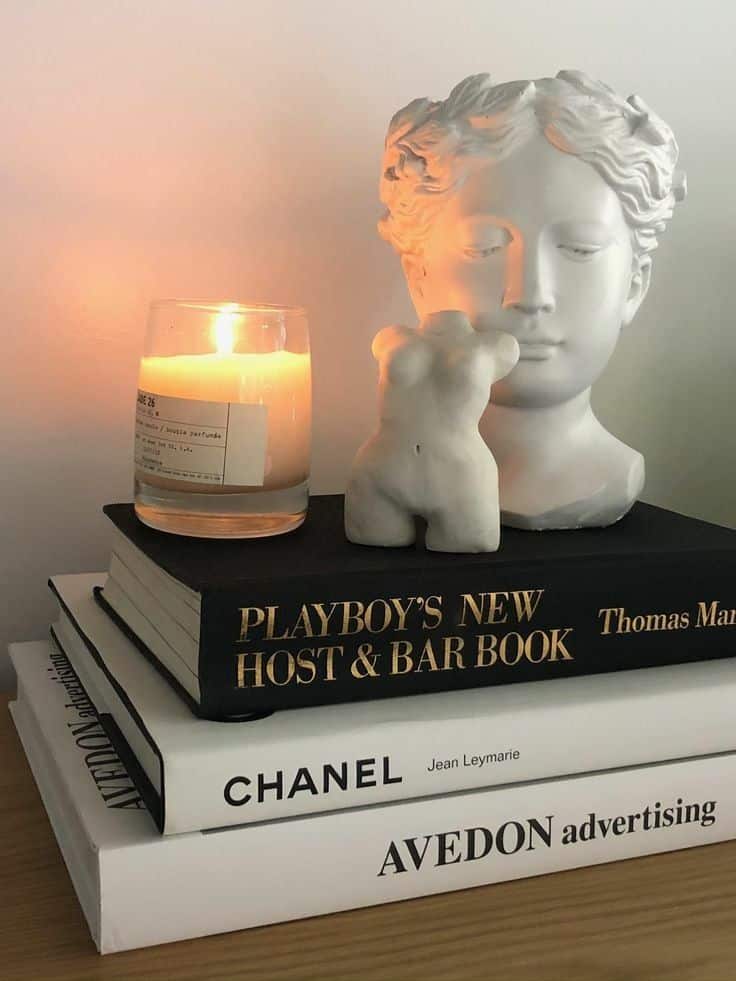 Decorative book stack with statue and candle, featuring titles like CHANEL and AVEDON advertising on a wooden surface.