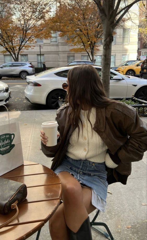 Woman in a brown jacket sits at a cafe table with a coffee cup, surrounded by autumn trees and parked cars.