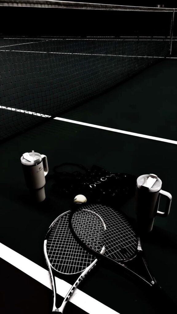 Tennis rackets, ball, and mugs on a dark court near the net, highlighting a sporty atmosphere.