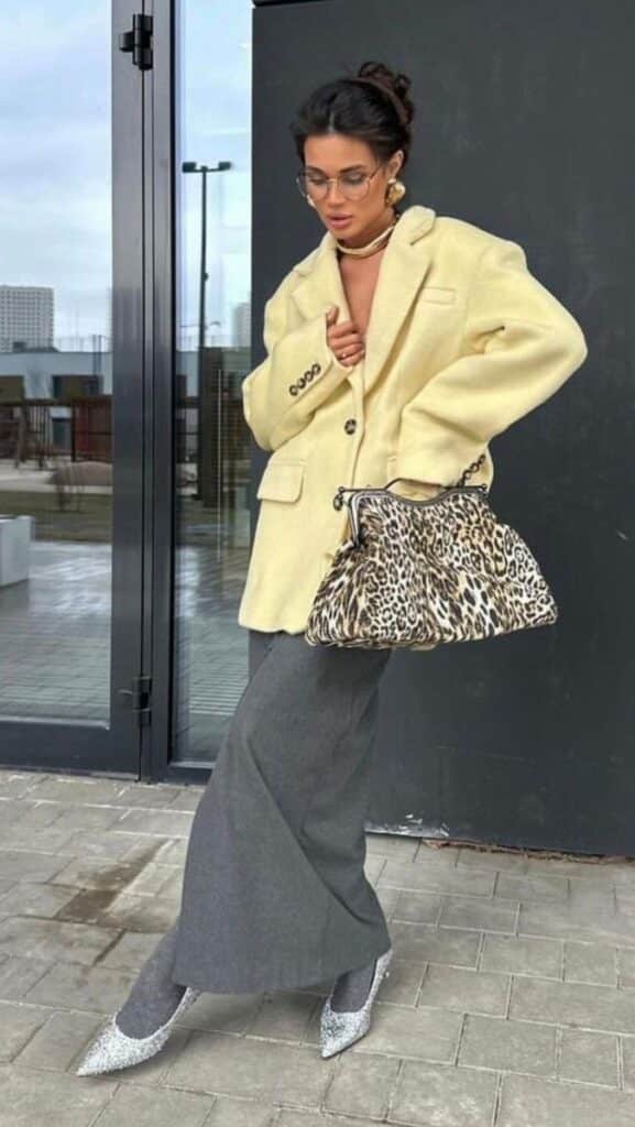 Woman in stylish oversized beige coat, gray pants, glitter shoes, holding leopard print bag. Urban fashion look.