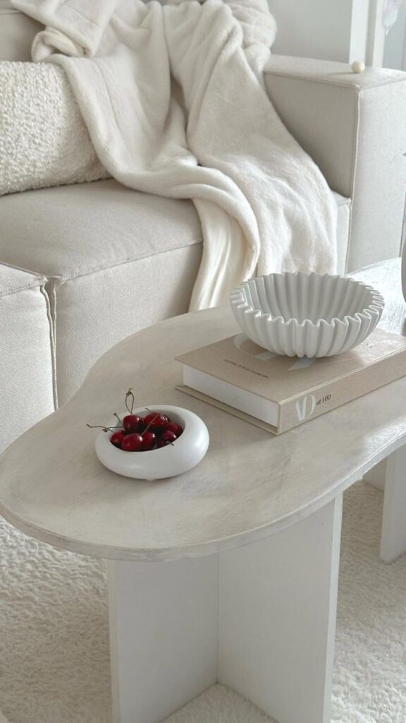 Cozy living room with a white sofa, draped blanket, modern table, decorative bowl, and cherries in a dish.