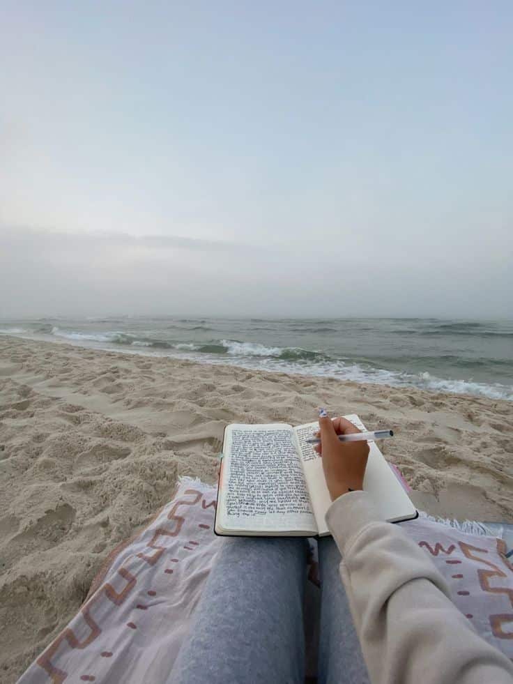 Person writing in journal by the beach, relaxing on a blanket with ocean waves in the background.