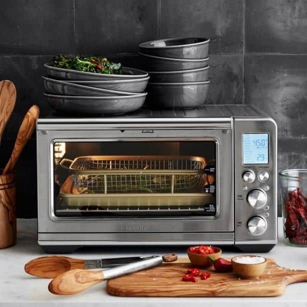 Stainless steel toaster oven on counter with bowls, spices, and wooden utensils; cooking in progress.