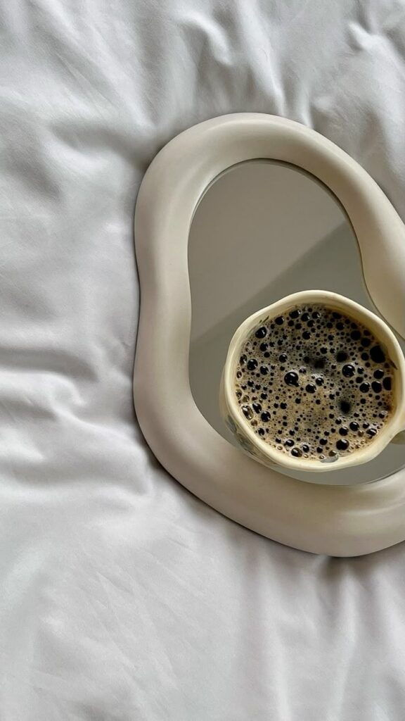 Coffee cup resting on an abstract mirror on a soft white fabric background, reflecting cozy morning vibes.