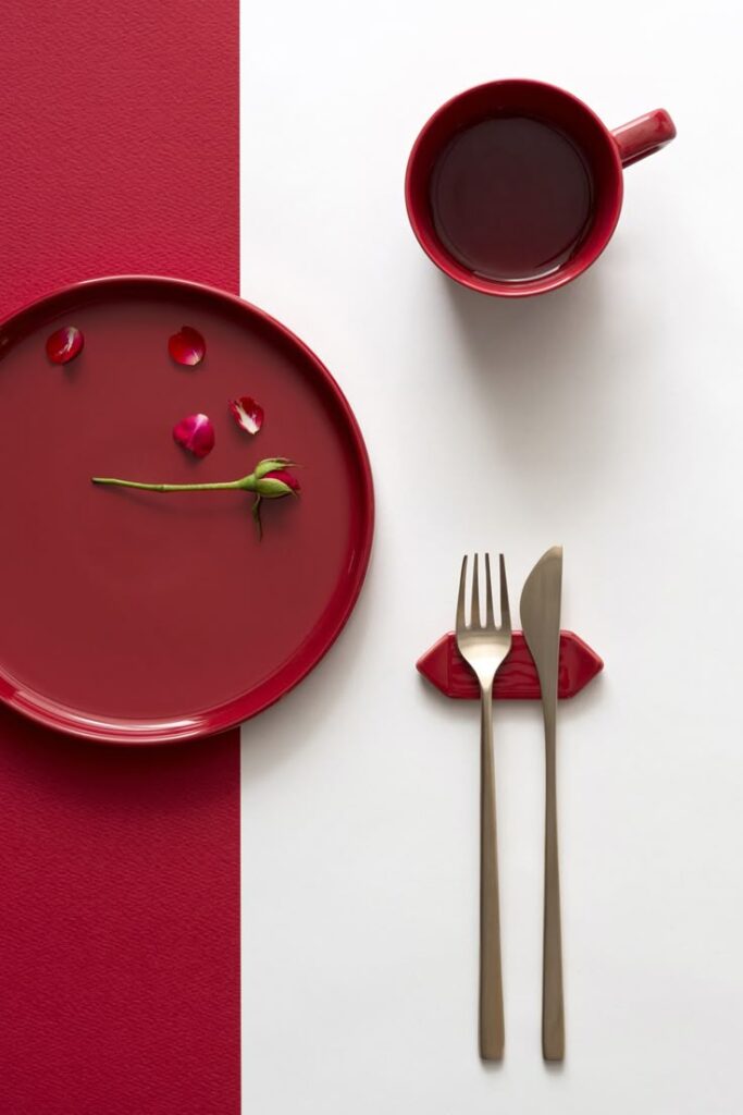 Red plate with rose petals and tableware on red and white background. Modern table setting with cup of dark liquid.