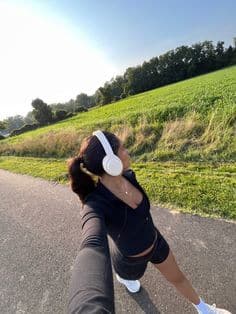 Woman jogging on a sunny day, wearing headphones and athletic wear on a rural path surrounded by greenery.