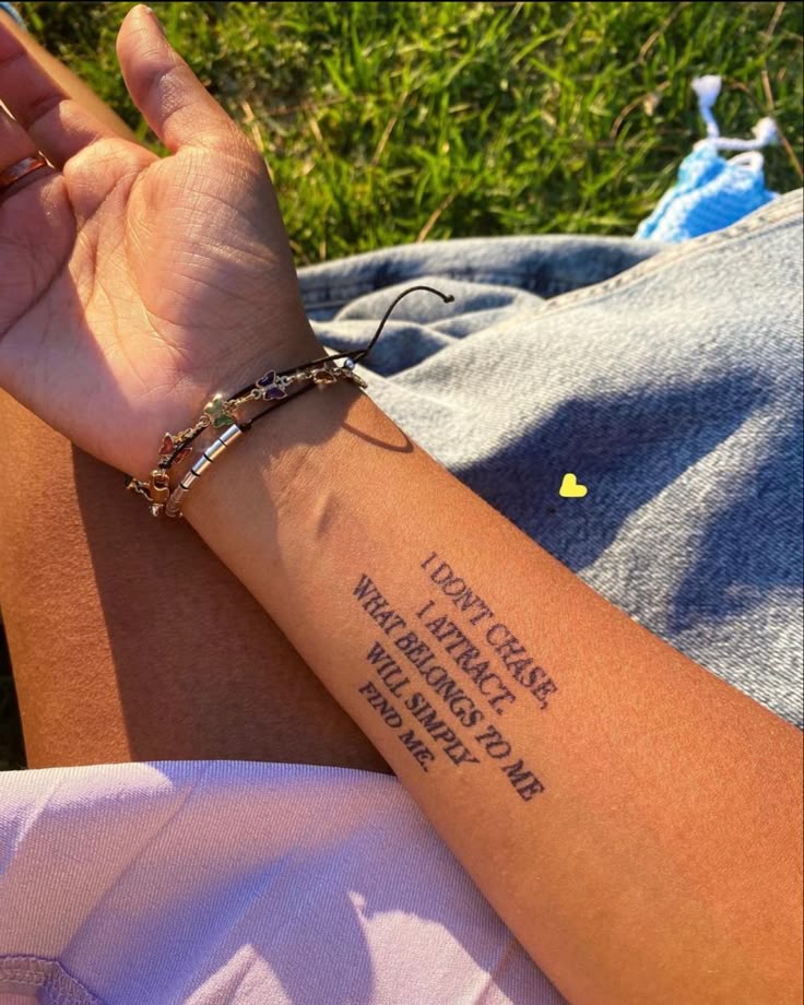 Arm tattoo quote about attraction, paired with stylish bracelets, resting on grass and denim.