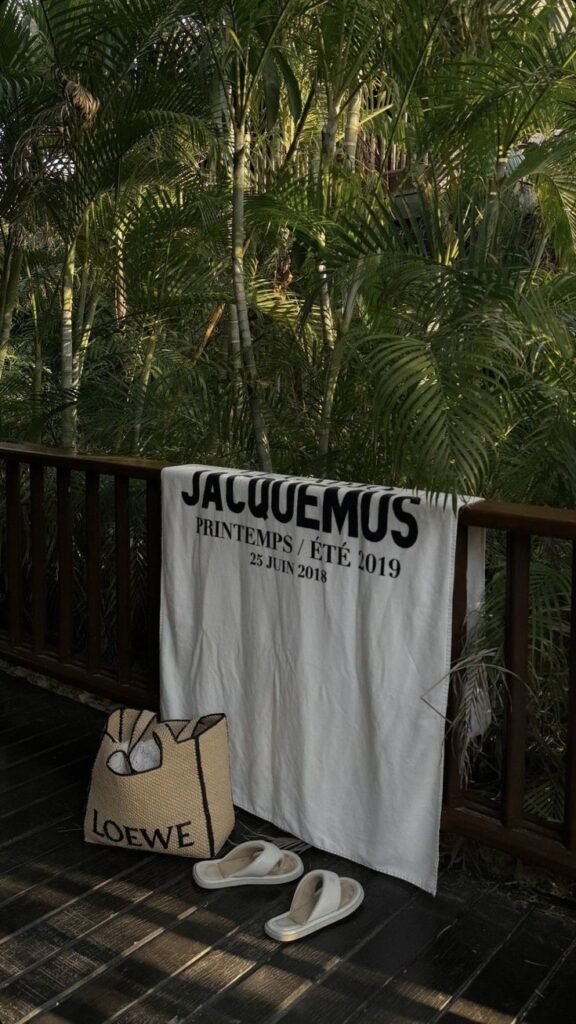 Tropical setting with Jacquemus towel, Loewe bag, and sandals on a wooden deck surrounded by lush greenery.