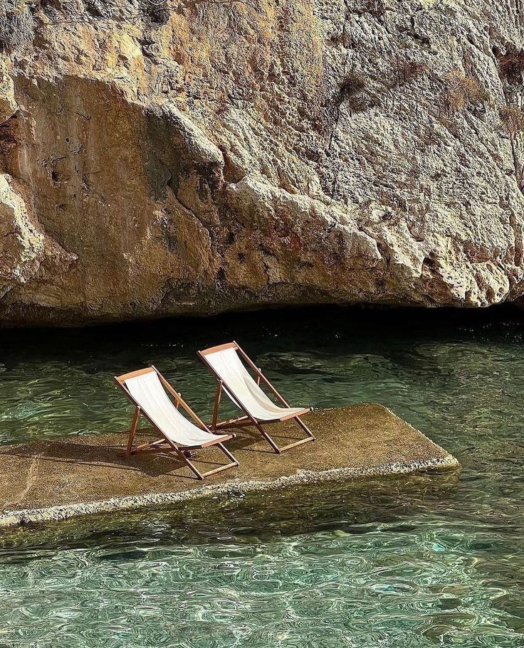 Two deck chairs on a stone platform by clear water and rocky cliff, serene sunbathing spot.