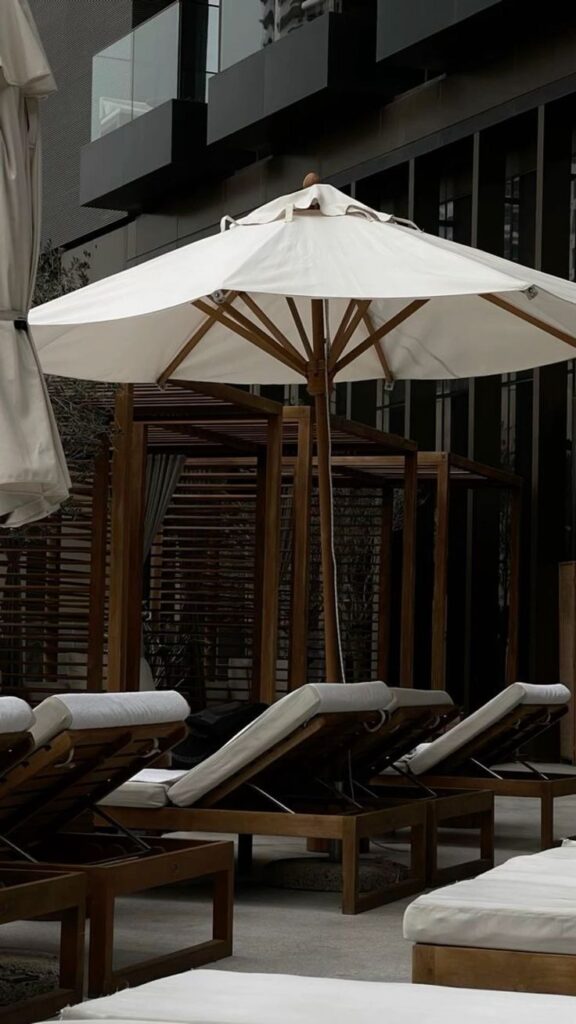 Outdoor patio with wooden loungers and a large white umbrella near a modern building. Tranquil relaxation spot.