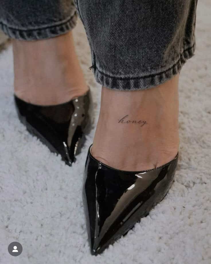 Close-up of black pointed heels on white carpet, showcasing ankle tattoo with the word honey.