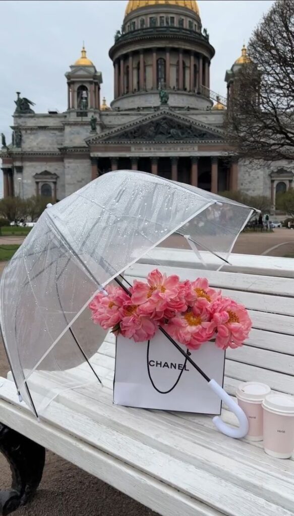Transparent umbrella and pink flowers in Chanel bag on bench with historic building backdrop.