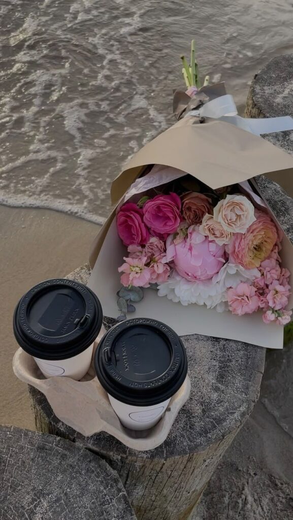 Bouquet of pink flowers and two coffee cups by the beach, resting on wooden posts.