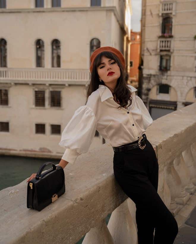 Fashionable woman in white blouse and black pants poses on a historic balcony, holding a black handbag.