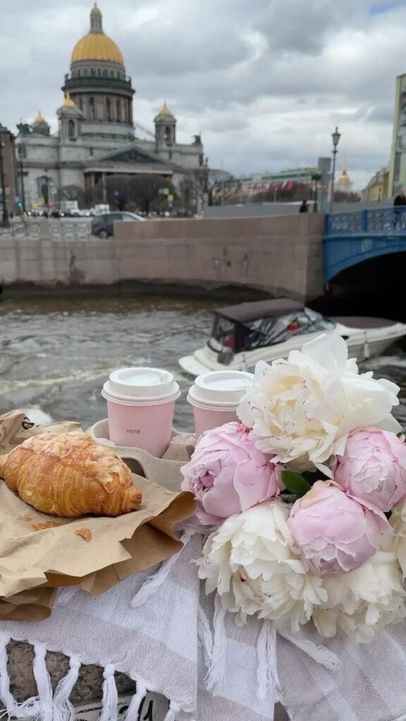 Croissant and coffee with pink flowers near a canal, cathedral in background, under a cloudy sky.